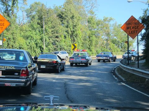 1991 Ford LTD Crown Victoria - convoy of police cars rounds a curve on a road past an orange LANE ENDS MERGE LEFT sign, seen through a windshield.