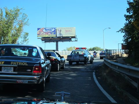 1991 Ford LTD Crown Victoria - black PATROL car and other cruisers travel a roadway ramp beneath a Tommy's Pizza billboard, photographed through a windshield.