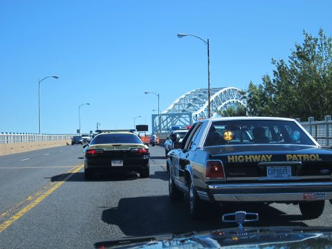 1991 Ford LTD Crown Victoria - black HIGHWAY PATROL sedan and other cruisers drive along a highway toward a steel arch bridge under clear blue sky.