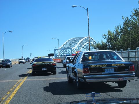 1991 Ford LTD Crown Victoria - black HIGHWAY PATROL sedan travels in a line of police cars on a highway approaching a steel arch bridge.