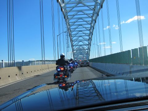 1991 Ford LTD Crown Victoria - Police motorcycles and a cruiser cross a long steel arch bridge, viewed over the hood of a following car.