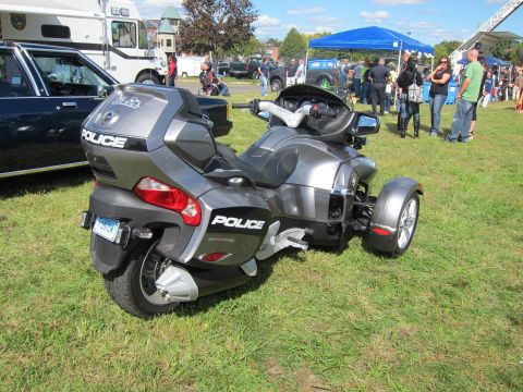 1991 Ford LTD Crown Victoria - gray three-wheeled POLICE Can-Am style motorcycle is displayed on grass at an outdoor show with tents and crowds behind.