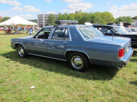 1991 Ford LTD Crown Victoria - light blue-gray Ford four-door sedan with a roof rack is parked on grass at a crowded outdoor car show.