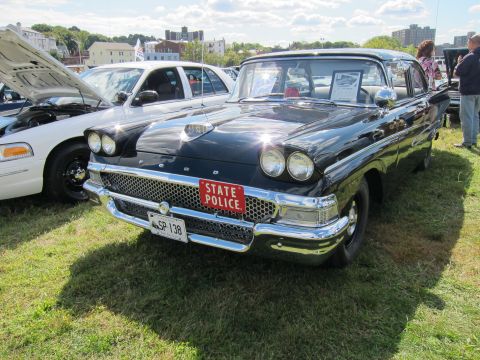 1991 Ford LTD Crown Victoria - black 1950s-era Ford sedan with a STATE POLICE front plate sits on grass at an outdoor car show beside a white cruiser.
