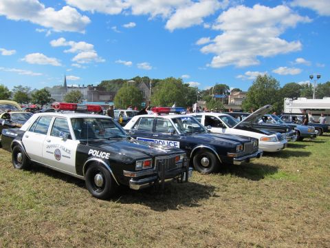 1991 Ford LTD Crown Victoria - row of marked police patrol cars, including black-and-white and dark blue sedans, lined up on grass at an outdoor show.