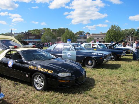 1991 Ford LTD Crown Victoria - black State Trooper coupe with gold lettering parked beside vintage marked state police sedans on grass.