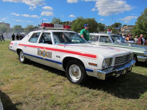 1991 Ford LTD Crown Victoria - white State Police sedan with a red side stripe and gold star door emblem parked on grass at a car show.