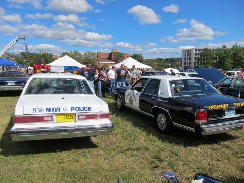 1991 Ford LTD Crown Victoria - white Miami Police sedan and a black-and-white Highway Patrol sedan parked rear-on to a crowd at an outdoor show.