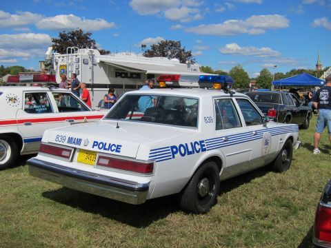1991 Ford LTD Crown Victoria - Rear three-quarter view of a white Miami Police sedan numbered 839 with blue stripes, parked on grass with crowd behind.