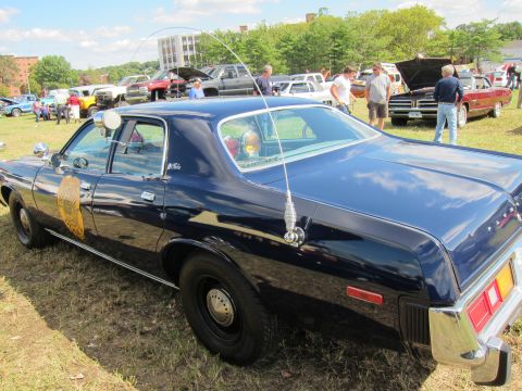 1991 Ford LTD Crown Victoria - dark blue four-door police cruiser with a tall whip antenna and gold door emblem is displayed on a grassy field among other classic cars.