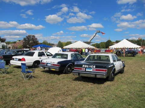 1991 Ford LTD Crown Victoria - Several white-and-black Police-marked sedans are parked side by side on grass with a crowd and event tents in the background.