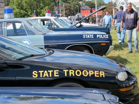 1991 Ford LTD Crown Victoria - black State Trooper coupe sits beside a blue State Police Plymouth Polara with its trunk open at an outdoor police car show.