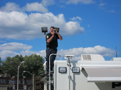 1991 Ford LTD Crown Victoria - man in a dark uniform stands atop a white command vehicle, raising a camera to photograph the scene against a blue sky.