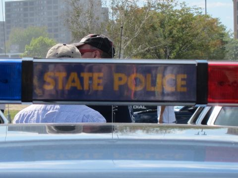 1991 Ford LTD Crown Victoria - close-up of a STATE POLICE light bar panel, with a man in a ball cap standing just behind it among parked cars.