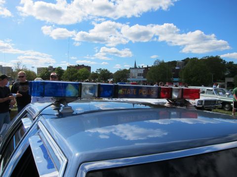 1991 Ford LTD Crown Victoria - State Police roof light bar tops a blue cruiser, with spectators and town buildings visible behind under a bright sky.