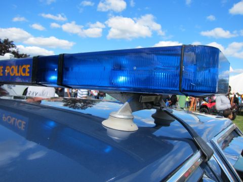 1991 Ford LTD Crown Victoria - blue police roof light bar is mounted on a cruiser roof, its lettering reflected on the paint, with a crowd and tents behind.