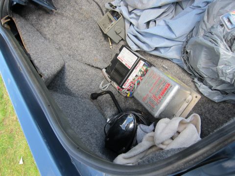 1991 Ford LTD Crown Victoria - Carpeted trunk interior of a blue car containing a sealed battery box, a wiring module, a folded car cover, and a helmet resting on a towel.