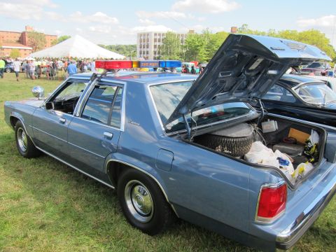 1991 Ford LTD Crown Victoria - Light blue four-door police sedan with a red-and-blue roof lightbar and open trunk, parked on grass at a sunny outdoor car show.
