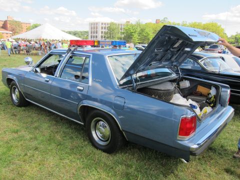 1991 Ford LTD Crown Victoria - Rear three-quarter view of a light blue four-door police sedan with a roof lightbar and open trunk, displayed on a grassy show field.
