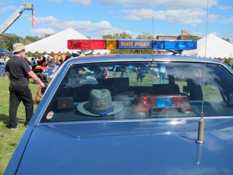 1991 Ford LTD Crown Victoria - Close view of a blue police car's roof showing an illuminated red-and-blue State Police lightbar and a trooper hat resting on the rear deck.