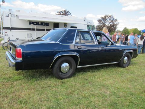 1991 Ford LTD Crown Victoria - Dark blue four-door sedan parked on grass in front of a white Police command/RV vehicle at an outdoor event with onlookers.