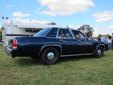 1991 Ford LTD Crown Victoria - Rear three-quarter view of a dark blue four-door sedan with blackwall tires, parked beside a white Police command truck on a show field.