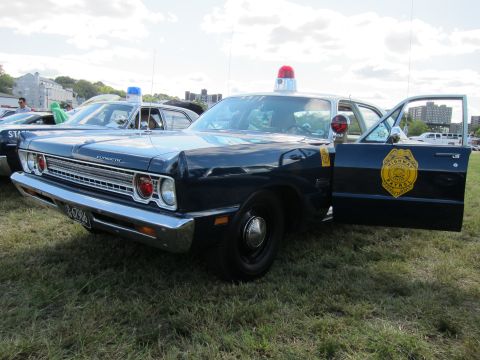 1991 Ford LTD Crown Victoria - Dark blue Plymouth police sedan with a single red roof beacon and a gold State Police shield on its open front door, shown on grass.
