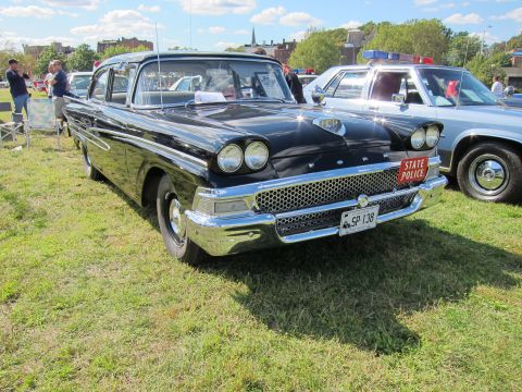 1991 Ford LTD Crown Victoria - Front three-quarter view of a black classic Ford sedan with quad headlights, FORD-lettered hood, and a red State Police plate at an outdoor…