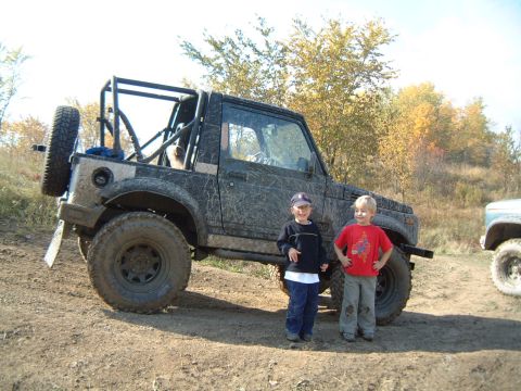 1986 Ford Bronco - A muddy Suzuki Samurai is parked off-road with two children posing in front of it, while another vehicle is visible to the right.