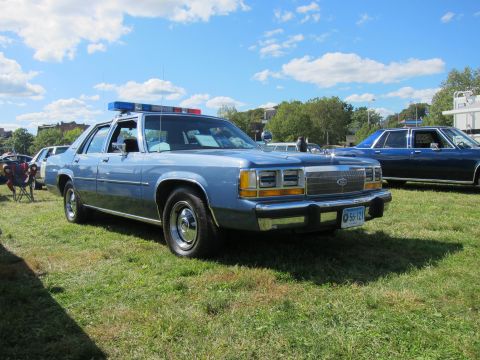 1991 Ford LTD Crown Victoria - light blue Ford LTD Crown Victoria with a red-and-blue lightbar parked on grass beside other vehicles at a show.