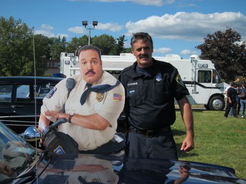 1991 Ford LTD Crown Victoria - uniformed officer with a mustache poses next to a cardboard cutout of a security guard leaning over a dark car hood.