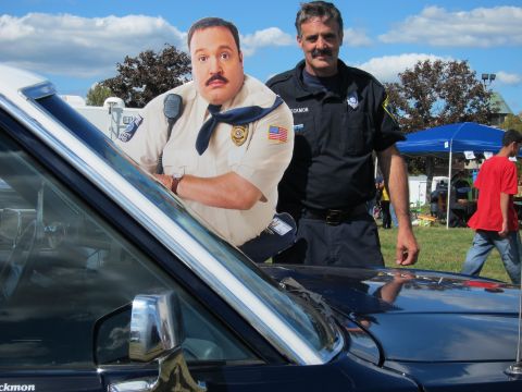 1991 Ford LTD Crown Victoria - mustached officer stands at the windshield of a dark sedan beside a cardboard cutout of a uniformed security guard.