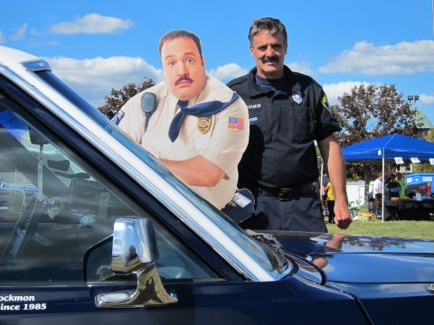 1991 Ford LTD Crown Victoria - uniformed officer stands beside a security-guard cardboard cutout over a dark sedan's hood under a blue sky.
