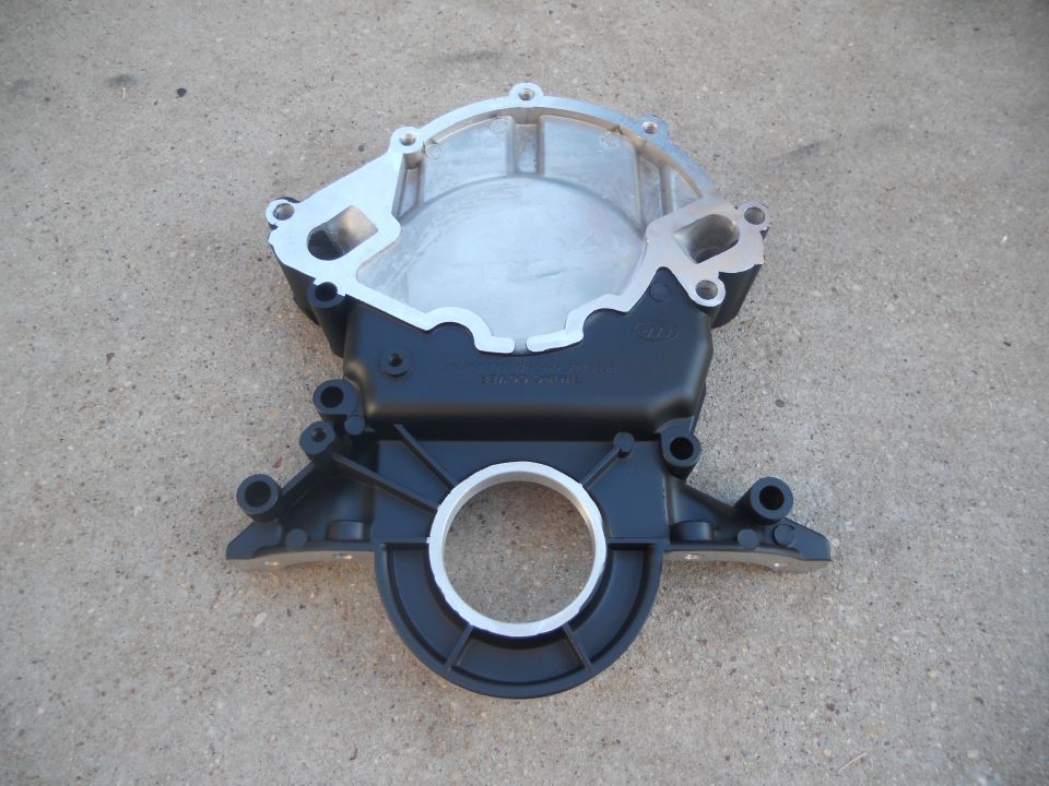 Engine timing cover part with aluminum and black plastic components, resting on concrete