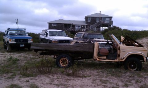 1979 Ford Bronco - Several trucks and a flatbed trailer are parked on a sandy area near a house on a cloudy day.