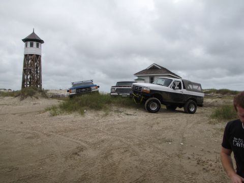 1979 Ford Bronco - Ford Bronco and two trucks parked on a sandy beach near a lighthouse and a house.