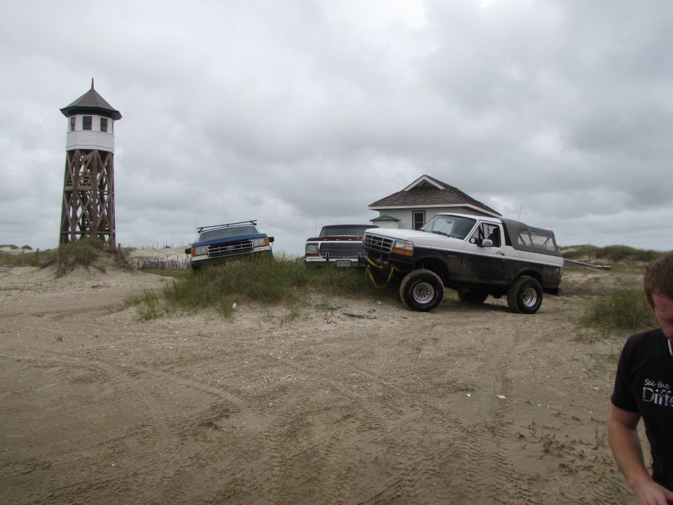 White and black Ford Bronco with a soft top, parked on sand near a house and a lighthouse. Two other trucks are visible in the background