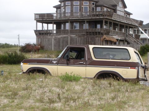 1979 Ford Bronco - A classic Ford Bronco is partially submerged in sand dunes, with a large beach house and a horse in the background.