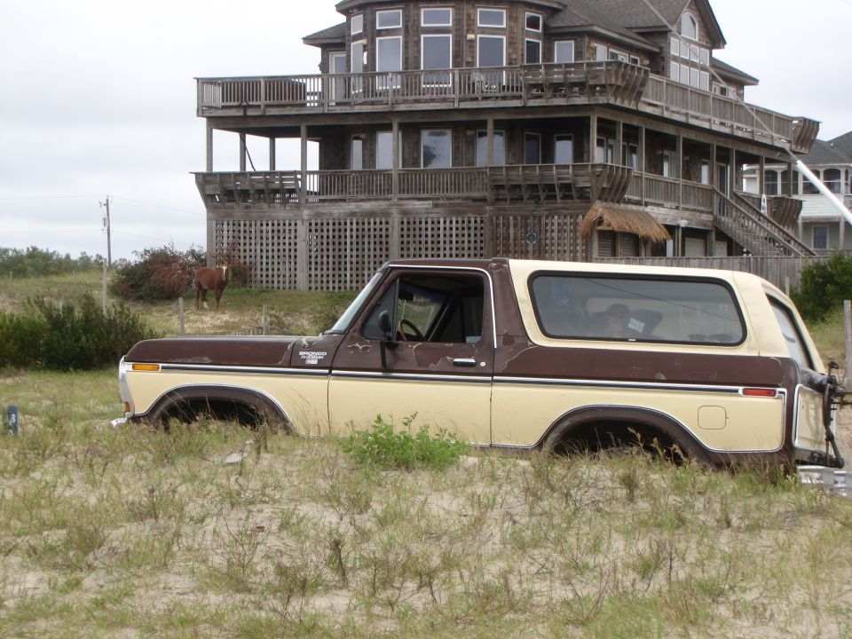 Brown and tan Ford Bronco stuck in sand dunes in front of a large beach house. A horse is visible in the background