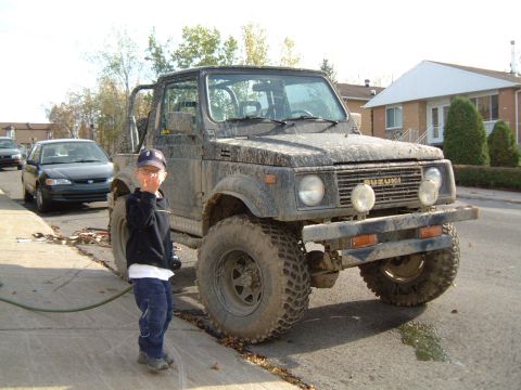 1986 Ford Bronco - A child stands next to a dirty Suzuki Samurai, with a residential street and houses visible in the background.