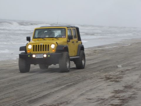1979 Ford Bronco - A yellow Jeep Wrangler drives along a sandy beach on an overcast day.