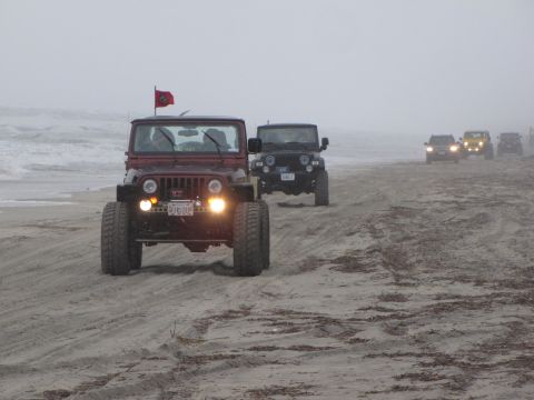 1979 Ford Bronco - A convoy of Jeeps, including a red Wrangler with a flag, cruises along a foggy beach.
