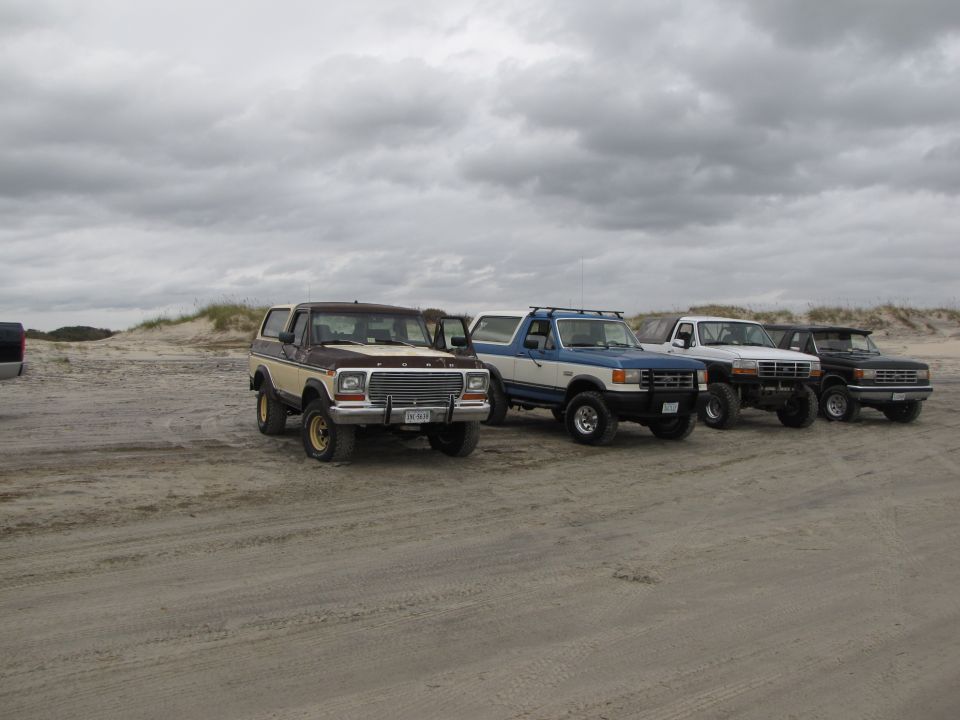Brown and tan Ford Bronco, blue and white Ford Bronco, white Ford F-150, and black Ford F-150 parked on a sandy beach under a cloudy sky