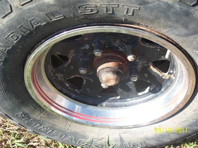 Close-up of a black wheel with a chrome lip and red stripe, mounted on a tire with visible text. A rusty hub is visible in the center