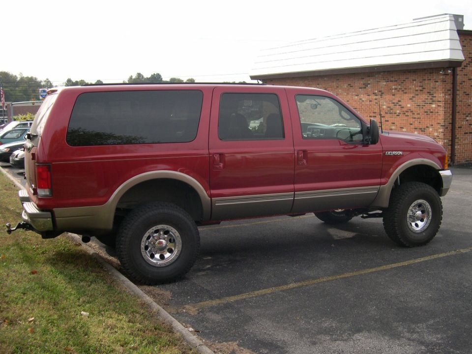Dark red lifted Ford Excursion SUV, side/rear view, oversized tires, parked by brick building