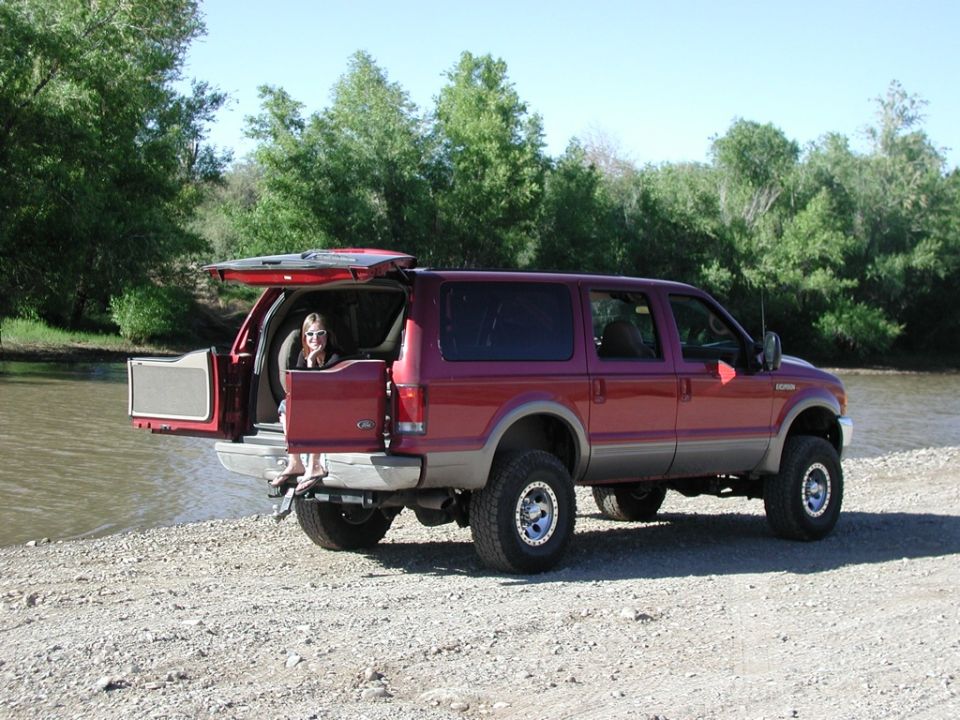 Maroon Ford Excursion SUV, rear hatch open, person sitting inside, parked by a river on gravel.