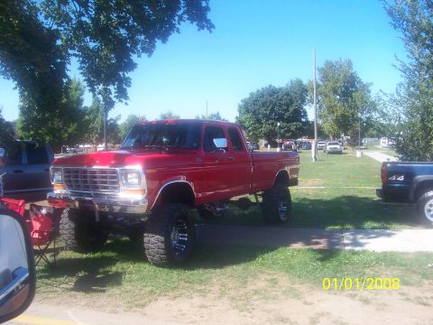 1989 Ford Bronco - A red pickup truck with a lifted suspension and large tires is parked on a grassy area next to other vehicles.