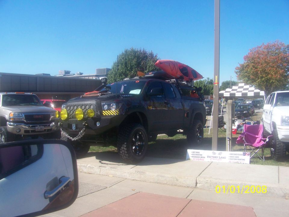 Modified black pickup truck with yellow fog lights, custom bumper, and kayak on roof rack. Another GMC truck is to the left. Date stamp reads 01/01/2008