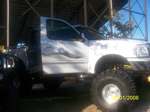 1989 Ford Bronco - A white pickup truck with custom graphics and oversized tires is shown, partially obscured by another vehicle. The photo is dated 02/01/200…