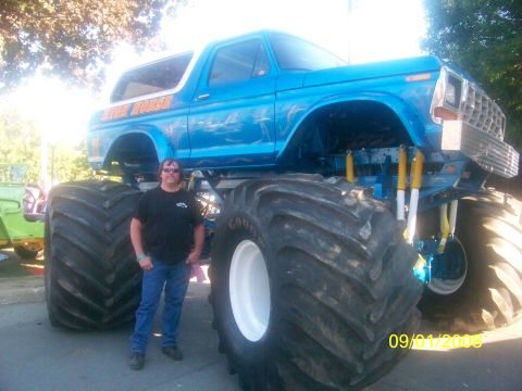 1989 Ford Bronco - A man stands beside a blue monster truck with 'High Horse' painted on the side, oversized tires and suspension.